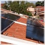 Foto de um telhado com paineis fotovoltaicos . Ao fundo o topo de uma árvore e uma paisagem urbana com muitas casas. Link no post sobre linhas de financiamento.