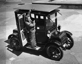 Foto de uma carro antigo com um homem em pé na porta do passageiro, que está aberta. Ele aponta para o teto do carro, no qual está um painel solar que cobre o carro em toda sua extensão. Foto de uma carro antigo com um homem em pé na porta do passageiro, que está aberta. Ele aponta para o teto do carro, no qual está um painel solar que cobre o carro em toda sua extensão.