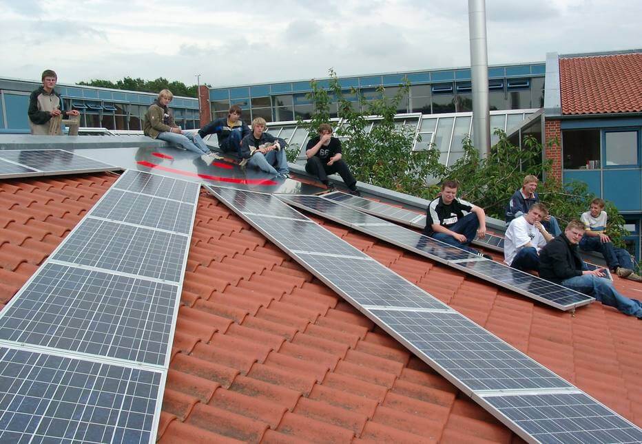 Foto de um telhado com painéis solarese alguns jovens sentados olhando para a câmera. A foto é de uma cidade da Alemanha que investiu em fontes renováveis para criar uma cidade verde.
