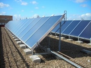 Conjunto de paineis solares térmicos instalados no solo com uma estrutura metálica. A foto foi tirada na diagonal da cena, na altura média de um observador. É possível ver duas fileiras de aproximadamente dez paineis cada. Ao fundo, céu azul. Foto no post de energia solar térmica e fotovoltaica.